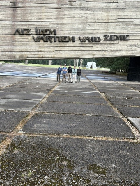 Members of the 2026 research group walking beneath the large Soviet-era entrance memorial at the Salaspils camp site.