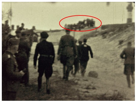 Historical Wiener-film still showing Jewish men being taken toward the burial trench while spectators stand in the distance, highlighted by a red oval.